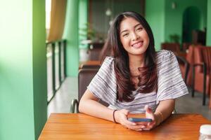 sonriente joven asiático mujer vistiendo vestir y sensación feliz, sentado a un restaurante para un desayuno mientras participación un teléfono inteligente foto