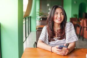 Smiling young Asian woman wearing dress and feeling happy, sitting at a restaurant for a breakfast while holding a smartphone photo