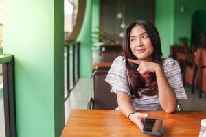 joven bonito asiático mujer sentado a un restaurante a tener desayuno mientras utilizando teléfono inteligente con dedo señalando a su Derecha lado foto