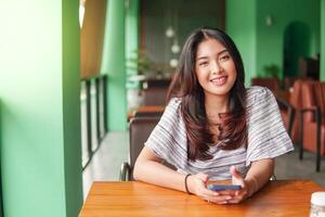 sonriente joven asiático mujer vistiendo vestir y sensación feliz, sentado a un restaurante para un desayuno mientras participación un teléfono inteligente foto