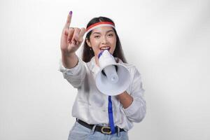 Energetic Indonesian woman in casual formal outfit wearing country flag headband while holding and shouting at megaphone, proudly showing little finger dipped in purple ink after voting for election photo