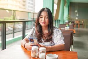 joven asiático mujer vistiendo vestir sentado a un restaurante para un desayuno, mirando a teléfono inteligente en un profundo pensamiento con grave cara foto