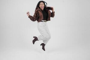 Active Asian girl is jumping and raising hands up wear brown jacket, isolated white color background photo