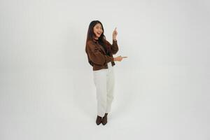 Smiling young Asian girl wearing brown jacket is standing and pointing to a copy space beside her. photo