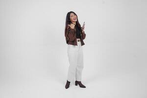 Smiling young Asian girl wearing brown jacket is standing while holding her phone and pointing to a copy space beside her. photo