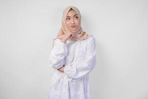 Thoughtful young Asian Muslim woman wearing white dress and hijab, holding her chin and thinking with serious expression over isolated white background photo