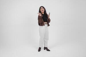 Smiling young Asian girl wearing brown jacket is standing while holding her phone and pointing to a copy space beside her. photo