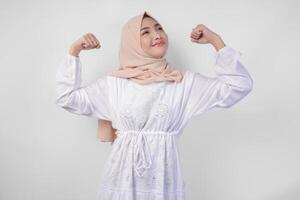 Excited Asian Muslim woman wearing white dress and hijab smiling confidently while posing strong gesture by lifting her arms and muscles. Ramadan and Eid Mubarak concept photo