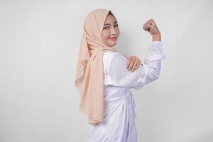 Excited Asian Muslim woman wearing white dress and hijab smiling confidently while posing strong gesture by lifting her arms and muscles. Ramadan and Eid Mubarak concept photo