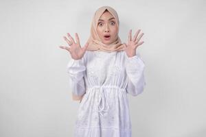 Portrait of young Asian Muslim woman wearing white dress and hijab with surprised expression and mouth wide open after hearing shocking news over isolated white background photo
