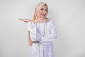 Excited Asian woman wearing hijab and white dress pointing to the copy space on her right side, standing over isolated white background. Ramadan and Eid Mubarak advertisement concept photo