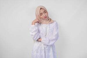 Thoughtful young Asian Muslim woman wearing white dress and hijab, holding her chin and thinking with serious expression over isolated white background photo