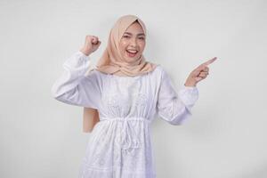 Overjoyed young Asian Muslim woman wearing hijab and white dress clenching fist winner gesture while pointing to the copy space beside her, isolated on white background photo