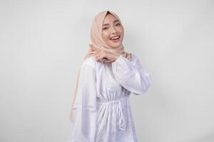 Cheerful Asian woman wearing hijab and white dress pointing to the copy space behind, standing over isolated white background. Ramadan and Eid Mubarak advertisement concept photo