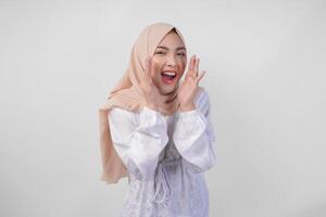 Beautiful young Asian Muslim woman in white dress and hijab shouting or announcing news, exciting event with hands on mouth, isolated on white background photo