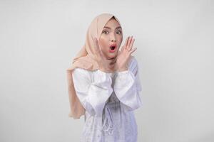 Beautiful young Asian Muslim woman in white dress and hijab shouting or announcing news, exciting event with hands on mouth, isolated on white background photo