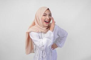 Beautiful young Asian Muslim woman in white dress and hijab shouting or announcing news, exciting event with hands on mouth, isolated on white background photo