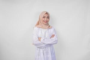 Confident young Asian Muslim woman wearing white dress and hijab smiling to the camera with arms crossed, standing over isolated white background photo