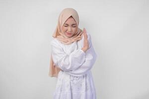 Young Asian Muslim woman wearing hijab and white dress gesturing a no rejection, refuse using hands to the copy space on left side, isolated by white background. Ramadan and Eid Mubarak concept photo