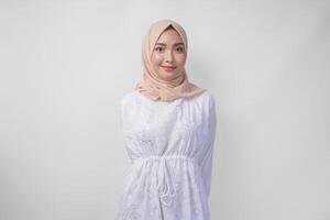 Attractive Asian Muslim woman in white dress and hijab smiling to the camera while standing formally over isolated white background photo