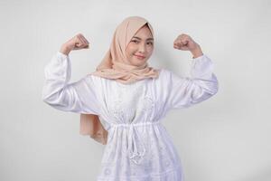 Excited Asian Muslim woman wearing white dress and hijab smiling confidently while posing strong gesture by lifting her arms and muscles. Ramadan and Eid Mubarak concept photo