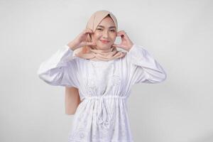 Young Asian Muslim woman in white dress and hijab making a heart love shape gesture with hands in various pose, isolated on white background photo