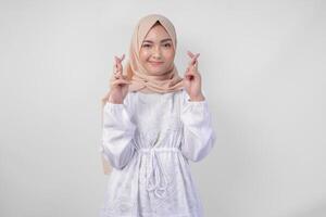 Beautiful Asian woman wearing hijab and white dress with fingers posing a wish me luck gesture, standing over isolated white background. Ramadan and Eid Mubarak concept photo