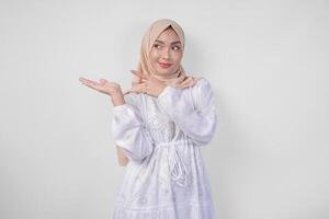 Smiling Asian woman wearing hijab and white dress pointing up to the copy space on her right side, standing over isolated white background. Ramadan and Eid Mubarak advertisement concept photo