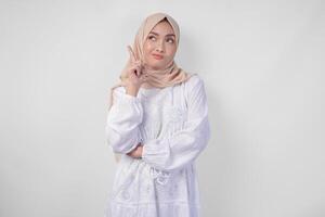 Thoughtful young Asian Muslim woman wearing white dress and hijab, holding her chin and thinking with serious expression over isolated white background photo