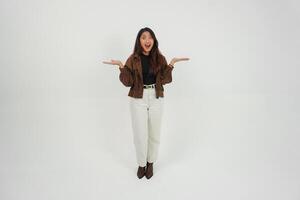 conmocionado joven asiático niña vistiendo marrón chaqueta es en pie y señalando a un Copiar espacio junto a su. foto