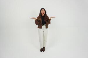 Shocked young Asian girl wearing brown jacket is standing and pointing to a copy space beside her. photo