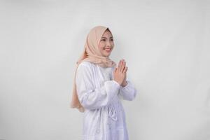 Beautiful Asian Muslim woman wearing white dress and hijab smiling while doing formal welcome or greeting gesture, standing over isolated white background photo
