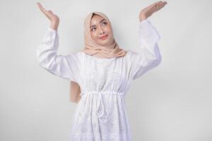 Happy Asian woman wearing hijab and white dress pointing up to the copy space above, standing over isolated white background. Ramadan and Eid Mubarak advertisement concept photo