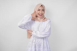 Young Asian Muslim woman wearing white dress and hijab smiling with happy expression while raising hand to do picking up phone gesture photo