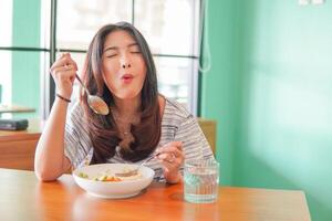 retrato de un emocionado joven asiático mujer vistiendo vestir sentado a un restaurante, comiendo y disfrutando desayuno con un alegre sonrisa foto