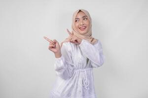 sonriente asiático mujer vistiendo hijab y blanco vestir señalando arriba a el Copiar espacio en su Derecha lado, en pie terminado aislado blanco antecedentes. Ramadán y eid Mubarak anuncio concepto foto