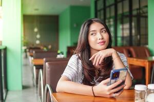 soñador joven asiático mujer vistiendo vestir sentado a un restaurante para un desayuno, participación un teléfono inteligente mientras apuntalando su barbilla perdido en pensamiento foto