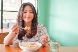retrato de un emocionado joven asiático mujer vistiendo vestir sentado a un restaurante, comiendo y disfrutando desayuno con un alegre sonrisa foto