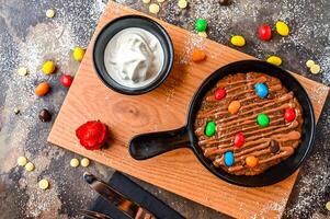 M and M Cookie Dough with mini chocolate candies, chocolate, whipped cream, knife and fork served in dish isolated on dark background closeup top view of cafe baked dessert food photo