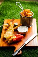 english Fish and Chips bucket served on wooden board with dip, knife and fork isolated on grassy background side view of fast food photo