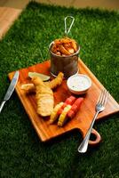 english Fish and Chips bucket served on wooden board with dip, knife and fork isolated on grassy background side view of fast food photo