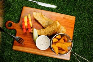 english Fish and Chips bucket served on wooden board with dip, knife and fork isolated on grassy background top view of fast food photo