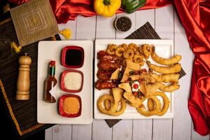 Assorted fried snacks platter with fried onion rings, shrimps, crackers, bq chicken wings, soy mayo dip sauce and black pepper served in dish isolated on napkin closeup top view on table italian food photo