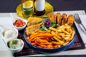 Snack platter include potato chips, french fries, fried mushroom, chicken wings and fish fry served in dish isolated on napkin side view of unhealthy food on table photo