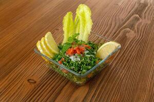 Tabbouleh salad with cucumber, tomato, onion, mint and lemon slice served in dish isolated on wooden table side view of arabic food photo