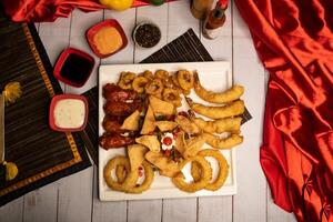 Assorted fried snacks platter with fried onion rings, shrimps, crackers, bq chicken wings, soy mayo dip sauce and black pepper served in dish isolated on napkin top view on wooden table italian food photo