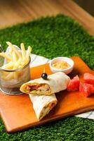 Chipotle Chicken Egg wrap roll filled with tomato, onion, cucumber, cheese with french fries, mayo dip and watermelon slice served on wooden board top view of fastfood appetizer photo