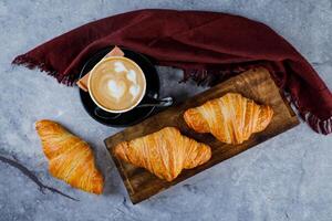 Plain Croissant served on wooden board with cup of coffee latte art isolated on yellow napkin top view of french breakfast baked food item on grey background photo