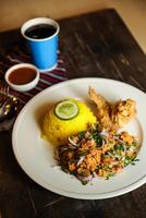 Hot Lemony Chicken Rice Meal served in plate with sauce, cold drink, spoon and fork isolated on wooden board side view of thai food photo