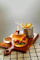 chipotle chicken burger include cheese slice, tomato, onion and lettuce leaf served on wooden board with dip and french fries bucket isolated on grey background side view of appetizer fast food photo